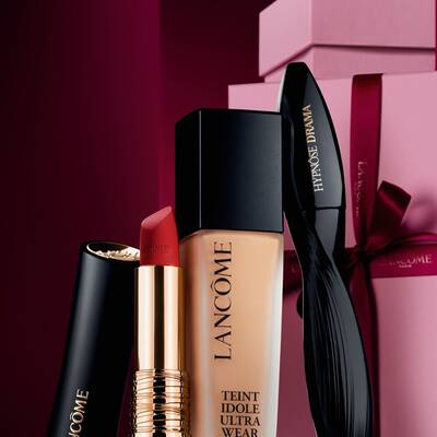 Una natura morta elegante composta dal rossetto opaco L'Absolu Rouge di Lancome, dal fondotinta Teint Idole Ultra Wear e dal mascara Hypnose Drama, su uno sfondo festivo rosso scuro e lussuoso.
