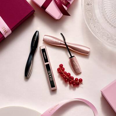 Una composizione flat-lay di prodotti trucco Lancome, con mascara e lucidalabbra, insieme a scatole regalo rosa, nastri di raso e un rametto di ribes rossi.