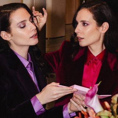 Due donne eleganti, che si rispecchiano a vicenda, indossano blazer di velluto e camicie di raso, mentre una si trucca sull'altra.