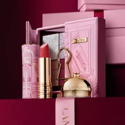 Un set regalo natalizio Lancome selezionato con rossetto, palette di ombretti e prodotti di bellezza dorati esposti su una custodia rosa decorativa.