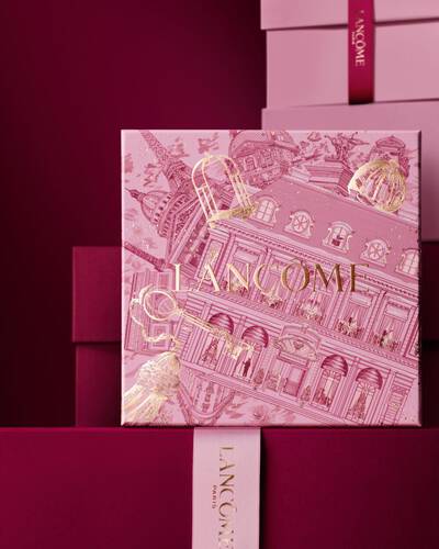 Immagine della scatola regalo rosa Lancome per le festivita davanti a una pila di scatole regalo rosa e rosso scuro, su uno sfondo rosso scuro.