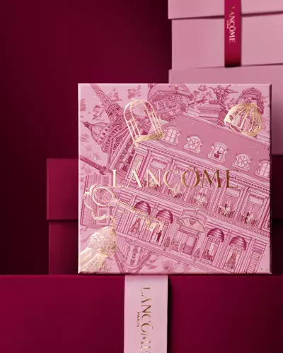 Immagine della scatola regalo rosa Lancome per le festivita davanti a una pila di scatole regalo rosa e rosso scuro, su uno sfondo rosso scuro.