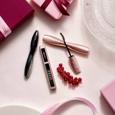 Una composizione flat-lay di prodotti trucco Lancome, con mascara e lucidalabbra, insieme a scatole regalo rosa, nastri di raso e un rametto di ribes rossi.