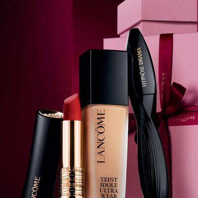 Una natura morta elegante composta dal rossetto opaco L'Absolu Rouge di Lancome, dal fondotinta Teint Idole Ultra Wear e dal mascara Hypnose Drama, su uno sfondo festivo rosso scuro e lussuoso.