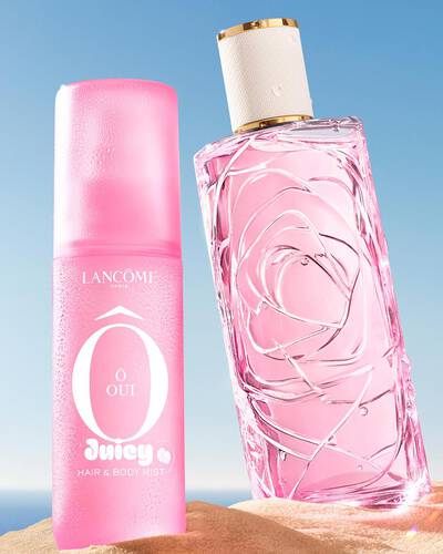 Tre bottiglie spray colorate, una gialla, una rosa e una verde, in piedi su una spiaggia sabbiosa accanto a un asciugamano a righe rosa. L'oceano e sullo sfondo sotto un cielo azzurro limpido.