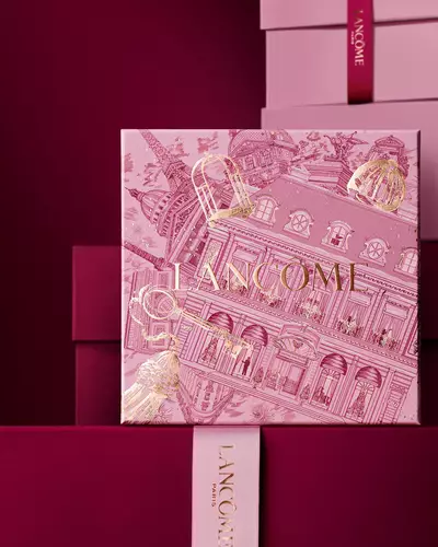 Immagine della scatola regalo rosa Lancome per le festivita davanti a una pila di scatole regalo rosa e rosso scuro, su uno sfondo rosso scuro.