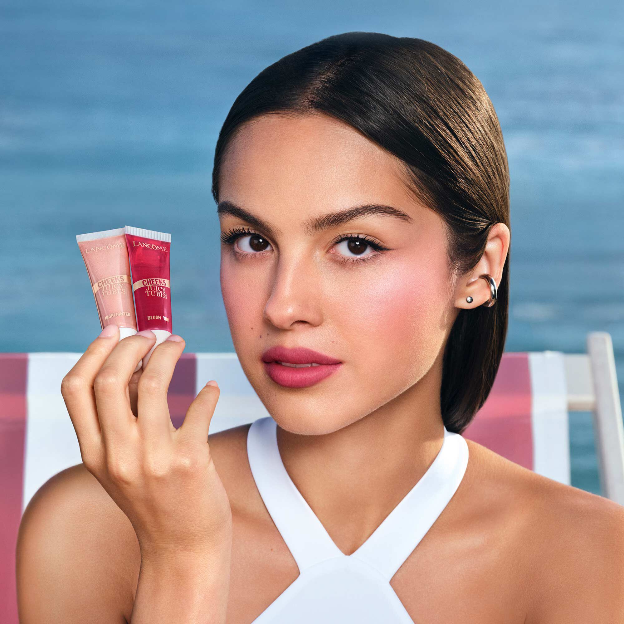 Olivia Rodrigo che tiene in mano un evidenziatore Juicy Tubes con uno sfondo da spiaggia.