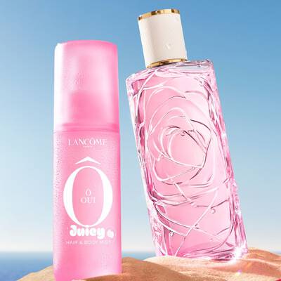 Una scena da spiaggia con tre coloriti flacchi di profumo in piedi sulla sabbia, uno rosa, uno giallo e uno verde, con un altro flacone rosa di profumo sdraiato su un fianco in primo piano. Un asciugamano a righe rosa e bianche e appoggiato sulla sabbia a sinistra. Lo sfondo mostra un cielo azzurro limpido che si trasforma in un oceano blu all'orizzonte.