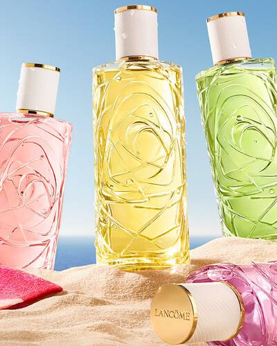 Due bottiglie rosa, una bottiglia di vetro trasparente profumato con un design texturizzato e una bottiglia spray, posizionata su una spiaggia sabbiosa. Un asciugamano da spiaggia rosa a righe piegato e accanto alle bottiglie. Sullo sfondo c'e un cielo azzurro limpido e l'oceano.