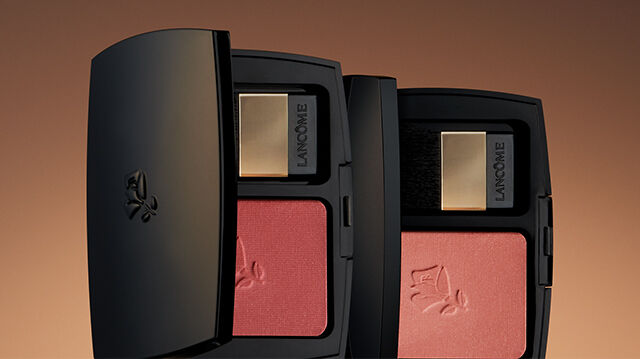Close-up of two sleek black cosmetic compacts containing blush, against a warm gradient background.