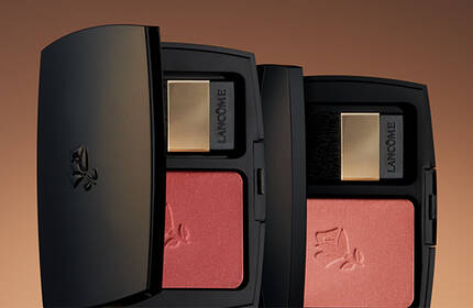 Close-up of two sleek black cosmetic compacts containing blush, against a warm gradient background.