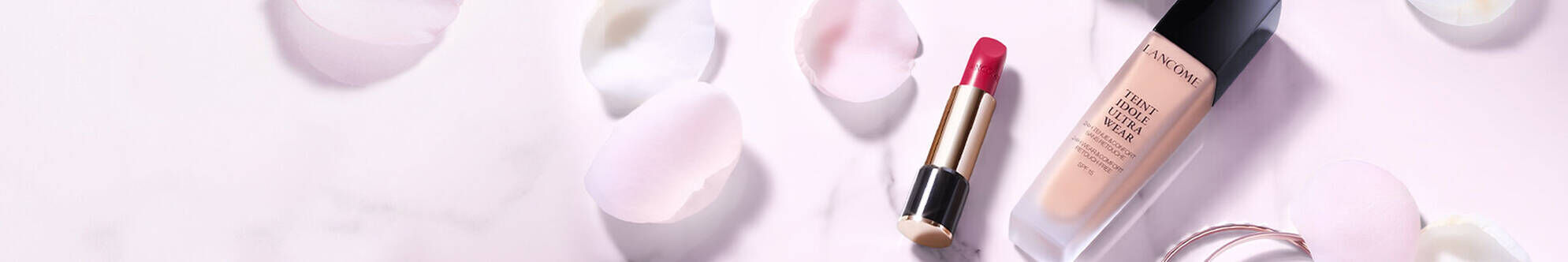 A bottle of foundation and a tube of bright red lipstick are elegantly placed on a soft pink background surrounded by scattered rose petals.