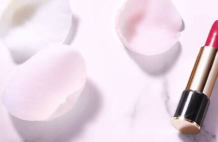 A bottle of foundation and a tube of bright red lipstick are elegantly placed on a soft pink background surrounded by scattered rose petals.