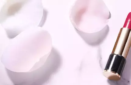 A bottle of foundation and a tube of bright red lipstick are elegantly placed on a soft pink background surrounded by scattered rose petals.