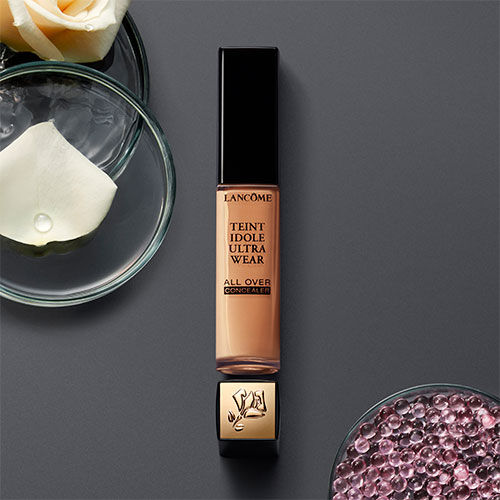 A bottle of Lancome Teint Idole Ultra Wear All Over Concealer sits on a dark surface, surrounded by glass bowls containing rose petals and decorative beads.