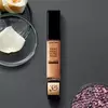 A bottle of Lancome Teint Idole Ultra Wear All Over Concealer sits on a dark surface, surrounded by glass bowls containing rose petals and decorative beads.