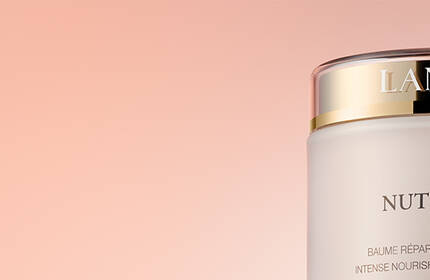 A close-up of a jar of Lancome Nutrix Royal Body cream, featuring elegant packaging with a gold lid against a soft pink background.