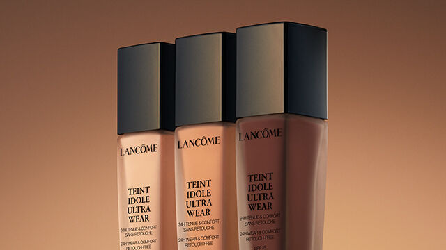 Three foundation bottles from Lancome displayed against a warm, gradient background.