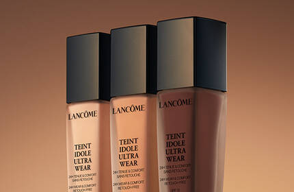 Three foundation bottles from Lancome displayed against a warm, gradient background.