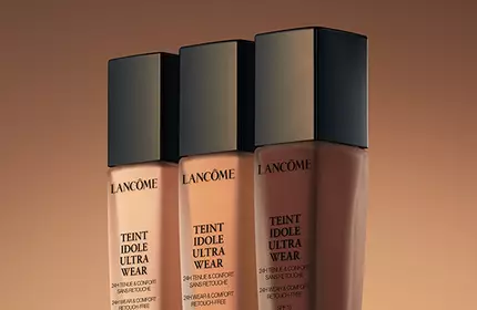 Three foundation bottles from Lancome displayed against a warm, gradient background.