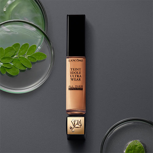 A bottle of Lancome Teint Idole Ultra Wear All Over Concealer placed on a dark surface, surrounded by small glass containers and green leaves.