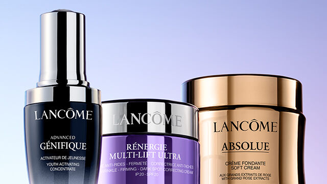 A close-up of three skincare products from Lancome, featuring sleek bottles with a modern design, set against a gradient background.