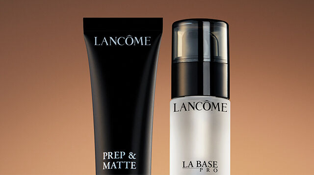 Two Lancome makeup products are displayed: a black tube labeled "Prep & Matte" and a clear bottle labeled "La Base Pro." Both are set against a warm, gradient background.