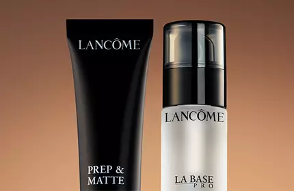 Two Lancome skincare products against a warm brown background: a black tube labeled "Prep & Matte" and a clear bottle labeled "La Base."
