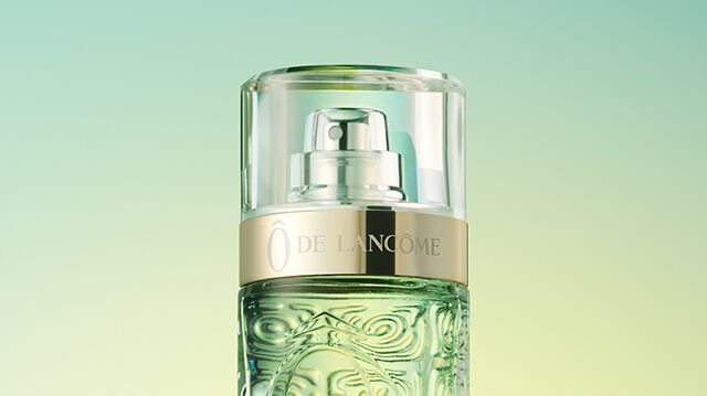 A close-up of a green glass perfume bottle with a silver and gold pump, set against a gradient background of soft green and yellow hues.