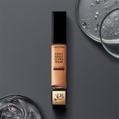 A tube of Lancome Teint Idole Ultra Wear All Over Concealer, placed on a dark surface beside circular glass containers filled with liquid.