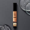 A tube of Lancome Teint Idole Ultra Wear All Over Concealer, placed on a dark surface beside circular glass containers filled with liquid.