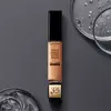 A tube of Lancome Teint Idole Ultra Wear All Over Concealer, placed on a dark surface beside circular glass containers filled with liquid.