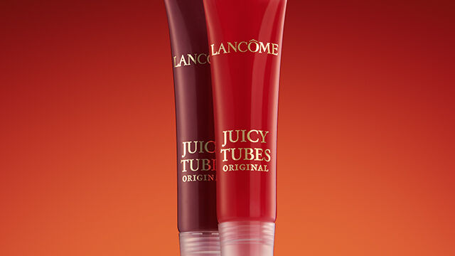 Two tubes of Lancome Juicy Tubes lip gloss standing upright against a gradient background of warm red tones.