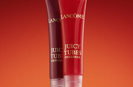 Two tubes of Lancome Juicy Tubes lip gloss standing upright against a gradient background of warm red tones.