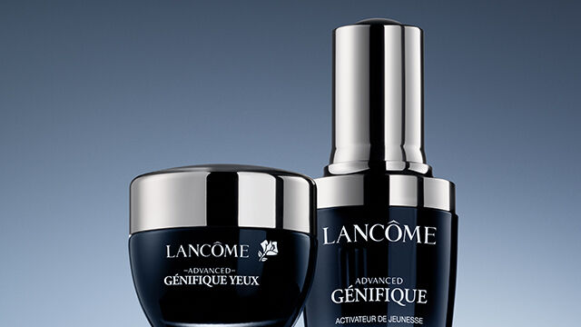 A visually appealing arrangement of two Lancome skincare products against a gradient blue background. The products include a small jar and a sleek serum bottle, both featuring elegant packaging.