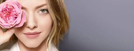 Ritratto di una giovane donna con capelli biondi, che tiene una rosa davanti all'occhio, sorridendo leggermente. Sullo sfondo, una tonalita di grigio chiaro.