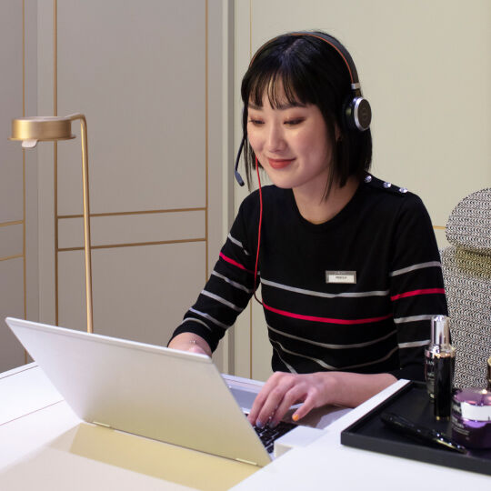 Una donna asiatica con cuffie lavora al computer portatile, seduta a un tavolo elegante, sorridendo mentre interagisce.