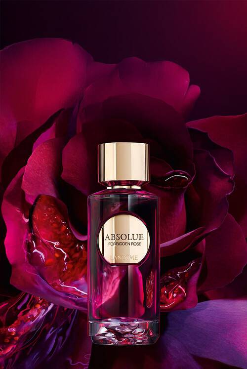 Una lussuosa bottiglia di profumo Lancome Absolue Forbidden Rose si trova davanti a petali di rosa dai toni rosso intenso e viola, con uno sfondo sfumato di rosso profondo. Il flacone presenta un tappo e un’etichetta dorati, creando un’immagine elegante e sofisticata.