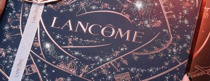 Scatola regalo di Lancome con un'elegante decorazione dorata e il testo "La vie est belle". Due mani la tengono in un ambiente suggestivo.