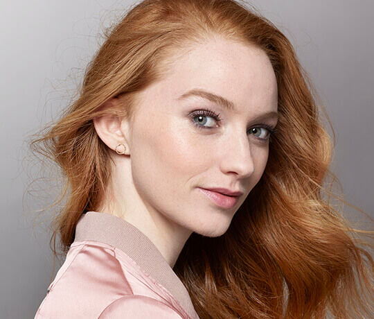Ritratto di una giovane donna con capelli rossi e lisci, che sorride con uno sguardo affascinante. Indossa un giubbotto di seta rosa e orecchini eleganti.