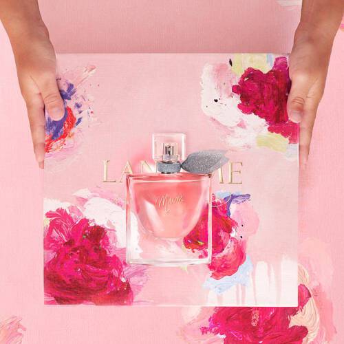 Una bottiglia di profumo 'Mimi' su uno sfondo rosa con fiori dipinti, tenuta da due mani.