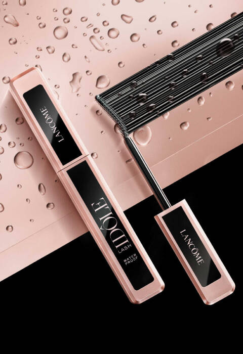 Un'immagine di un mascara Lancome con packaging rosa e un pettine per ciglia, posizionati su una superficie con gocce d'acqua.
