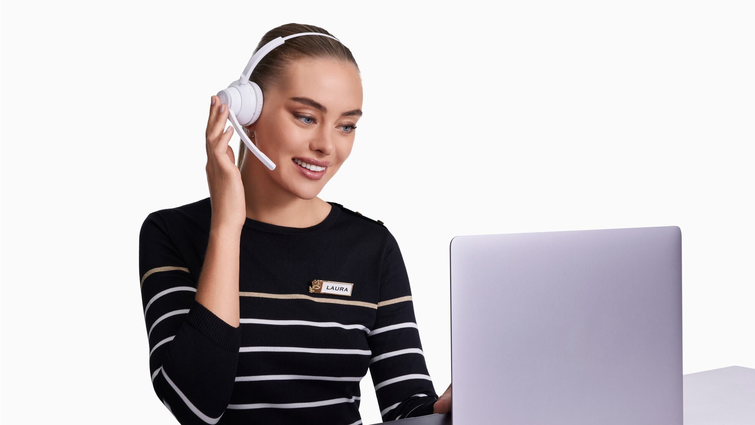 Una donna sorridente con cuffie bianche parla al microfono mentre guarda un laptop. Indossa un maglione a righe e ha un nome identificativo sulla maglietta.