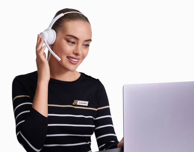 Una donna sorridente con cuffie bianche parla al microfono mentre guarda un laptop. Indossa un maglione a righe e ha un nome identificativo sulla maglietta.