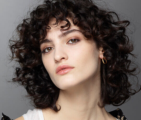 Ritratto di una giovane donna con capelli ricci e scuri, indossa orecchini dorati e ha un'espressione serena.