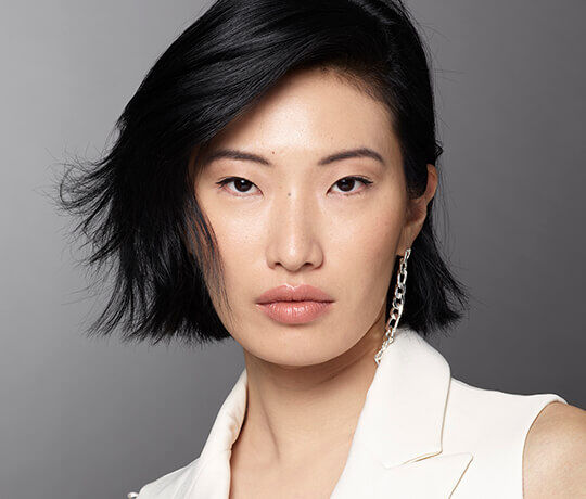 Ritratto di una donna con capelli neri e corti, che indossa un blazer bianco e orecchini a catena, con uno sguardo intenso.