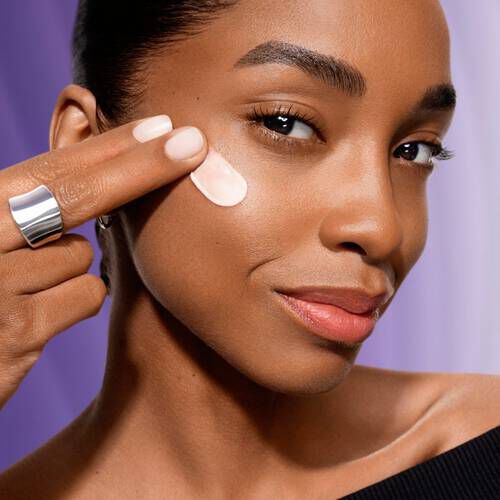 Persona applica una crema viso sul viso, sorridendo, con uno sfondo viola sfumato.