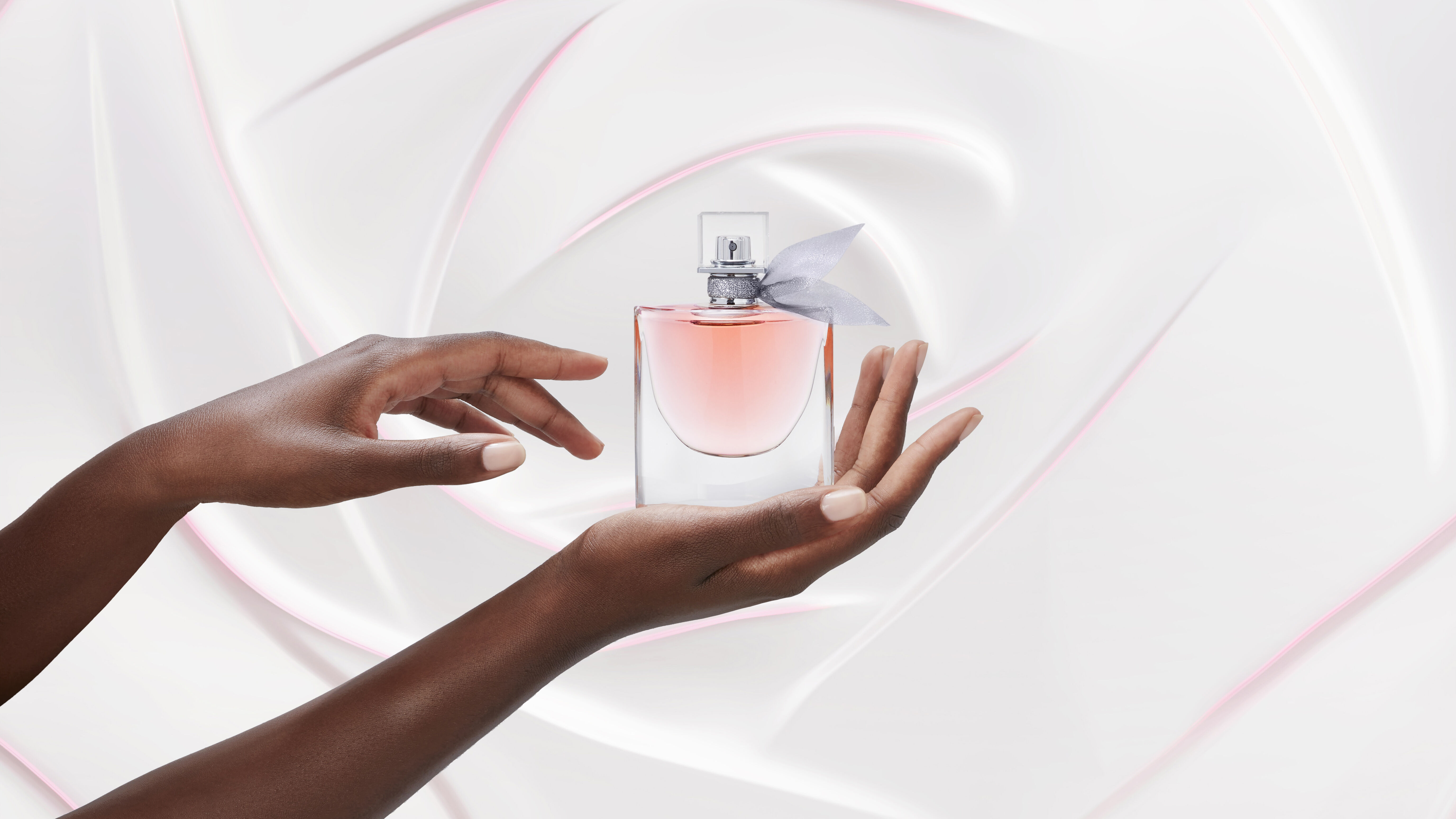 Una mano che tiene un flacone di profumo con un tocco di rosa, posato su uno sfondo elegante e lievemente ondulato.