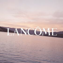 Testo alternativo: Logo di Lancome su uno sfondo di un lago al tramonto, con colori tenui nel cielo e nelle acque.