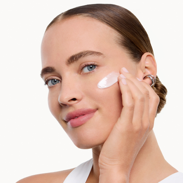 Donna che applica una crema viso rosata sulla guancia, pelle luminosa e sfondo bianco pulito.